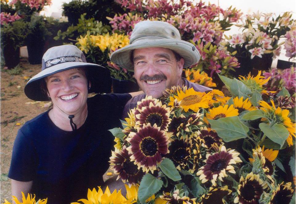 couple nestled in sunflower patch wearing hats and smiling towards the camera