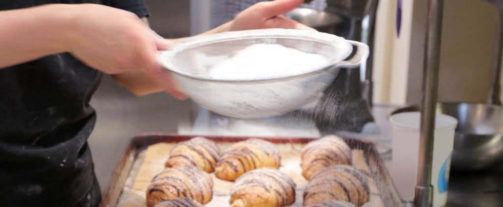 side view of baker sifting white powder over chocolate croissants wearing black jacket
