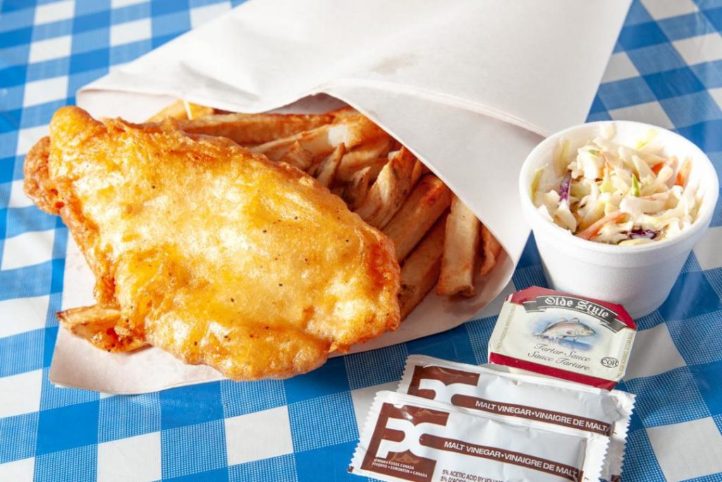 top view of battered fish and chips on blue and white checkerboard tablecloth with ramekin coleslaw and ketchup packs to the right