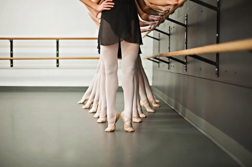 bottom view of ballerinas all with different foot positions wearing slippers holding a bar