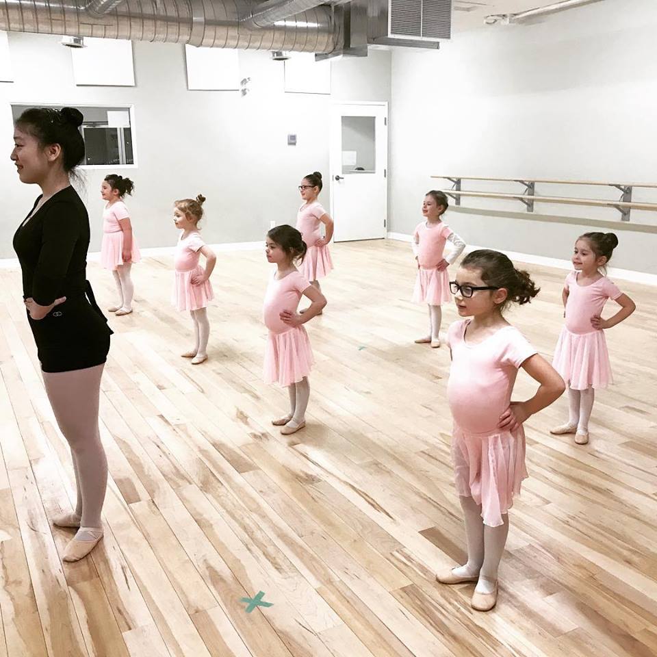 older woman in black teaching young dancers in pink dresses all looking towards mirror with hands on hips
