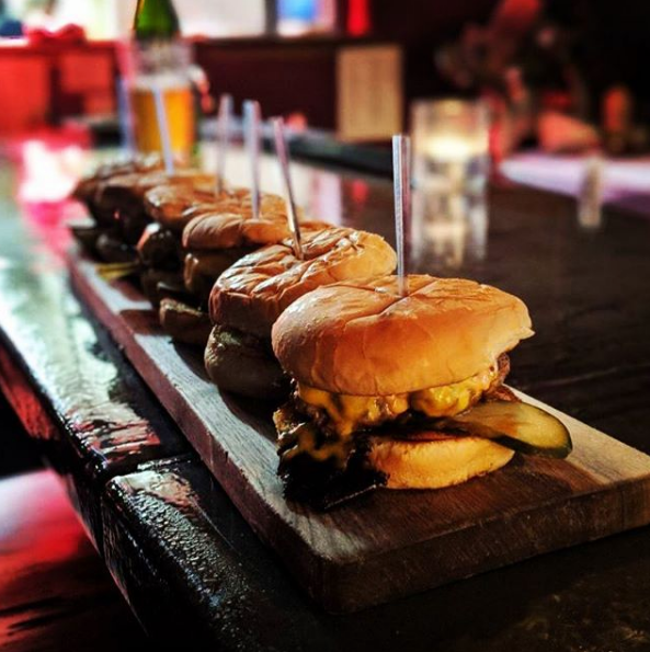 side view of paddle of mini burger sliders with garnish on wooden plank on black bar top