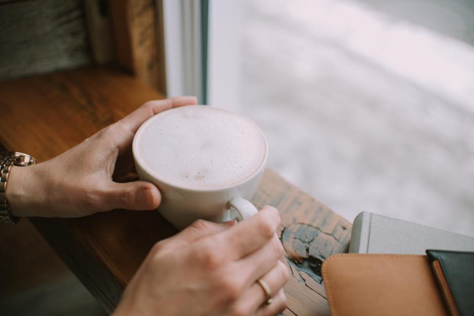 close up of hands holding frothy coffee cup in front of window on a wooden table