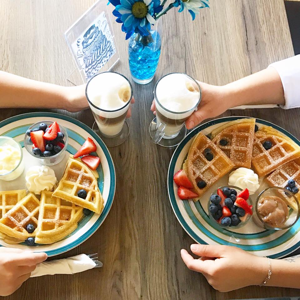 overview of two peoples waffle plates with berry garnish and lattes in hand on a wooden table