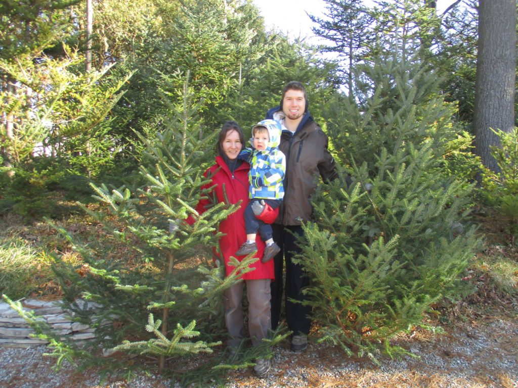 mom and dad holding young child with christmas trees in the background smiling at camera