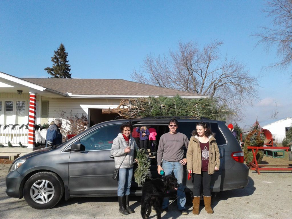 family posing in front of van with christmas tree tied on top on a nice day