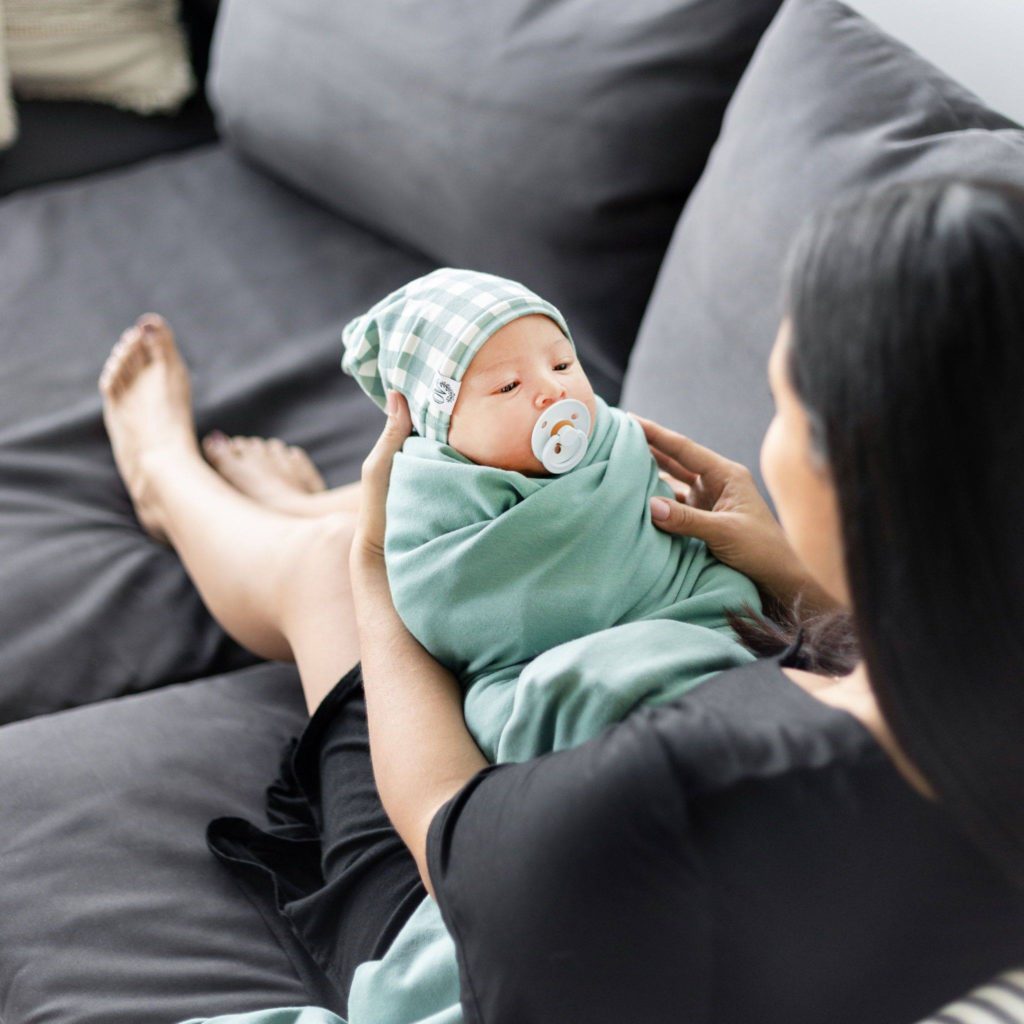 woman sitting on couch with knees holding a very young baby wrapped in teal swaddle