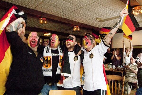 four men linking arms wearing german face paint, jerseys and holding flags yelling past the camera