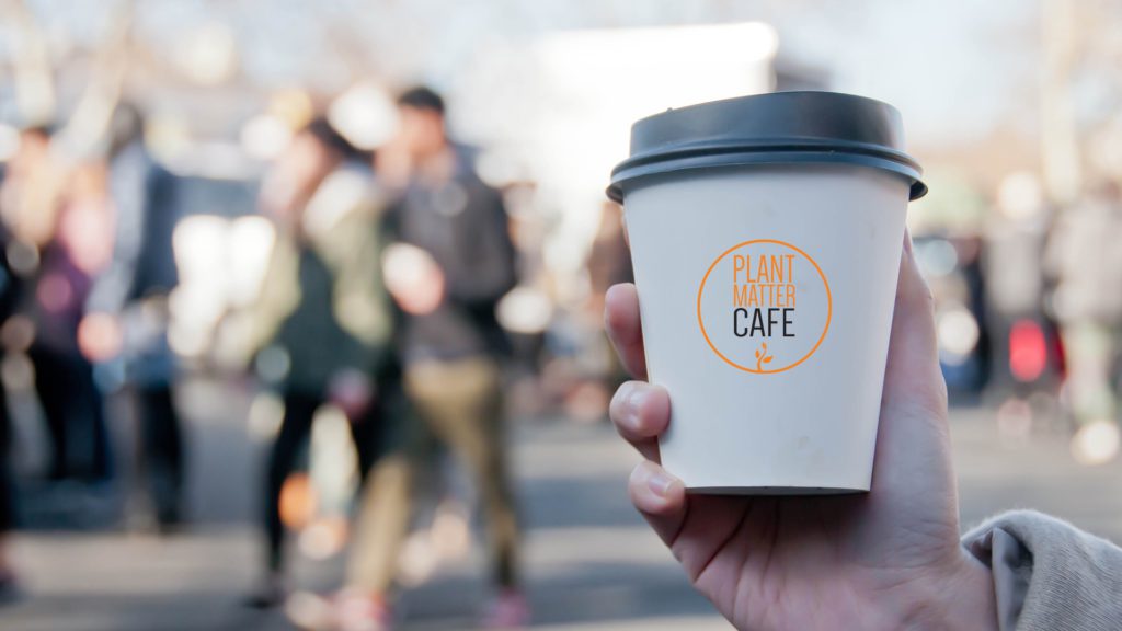 man holding to go small coffee cup in front of crowd with people blurred and focus on cup