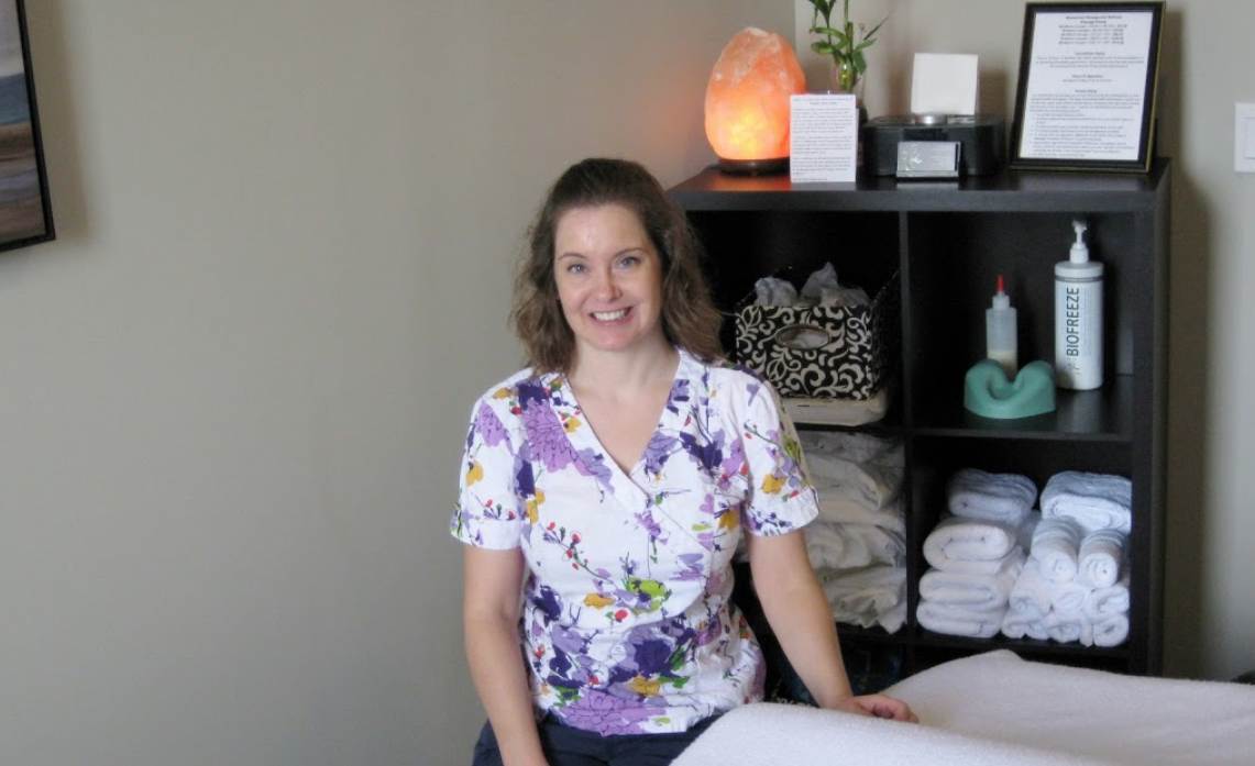 woman sitting beside a massage table wearing scrubs amongst a salt lamp and lotion sitting on a shelf