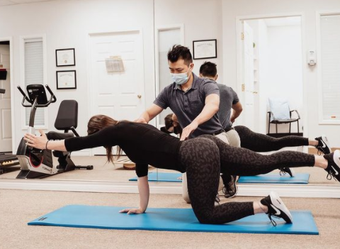 woman stretching on a yoga mat with an instructor with a mask helping her shape