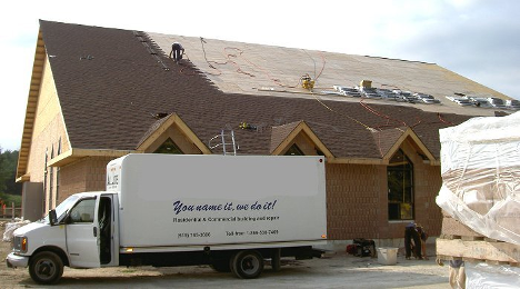 large building under construction with shingle bags on the roof and a large truck parked in front