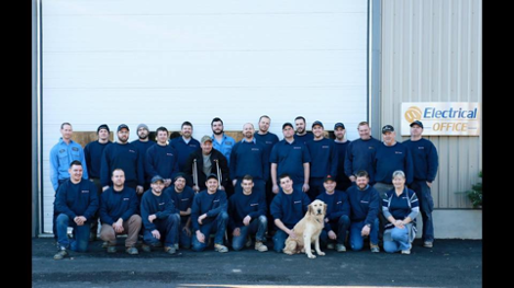 many employees standing and kneeling outside a warehouse all wearing matching blue shirts and a dog in the front middle