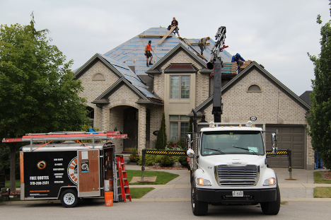 many men replacing the shingles on a roof with a large truck in the driveway and a trailer parked on the road