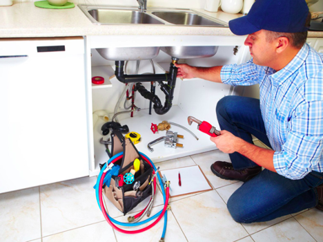 man in a checkered shirt and blue hat working on the pipe under a kitchen sink with a toolbag beside him