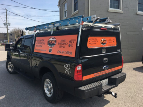 a black and orange work truck with company name and phone number on the side with a ladder on the roof parked outside of a building