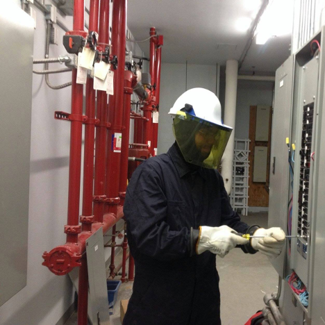 man wearing protective head gear and gloves using a screwdriver on a fuse box in a warehouse