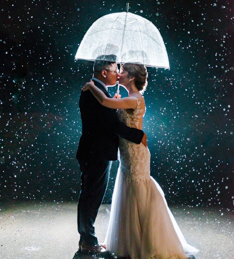 Bride and groom holding umbrella kissing under the rain