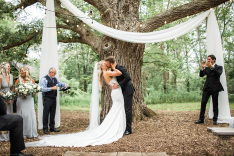 Bride and groom standing under tree with lights and linen kissing with bridal party to the left