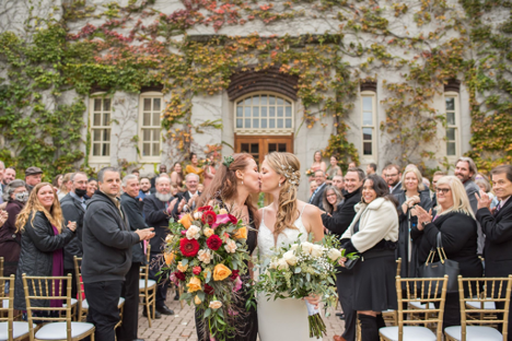 two women in aisle kissing holding bouquets surrounded by family and friends