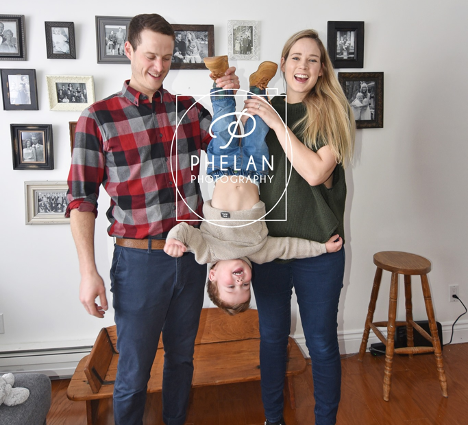 two parents holding young boy upside down smiling with photo collage on the wall behind them