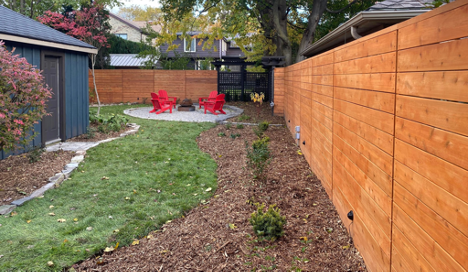 large wooden backyard fence with garden feature to the left, fire pit in the background and shed in the back