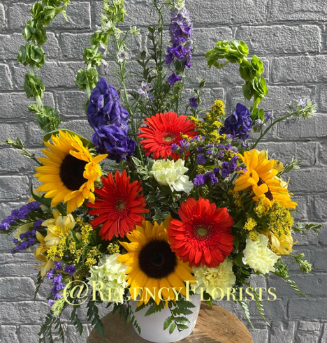 Colourful flower bouquet sitting in a white vase on a circular table with purple, red and yellow flowers against a brick wall