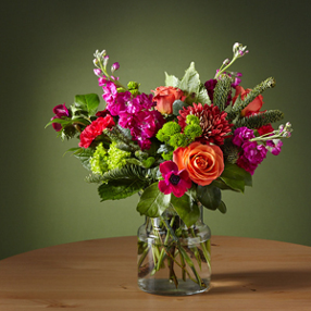 large flower bouquet in a glass vase sitting on a circular table with orange and pink flowers against a green wall