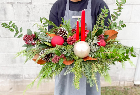 man wearing apron holding large christmas arrangement with pine, candles, christmas ornaments and pine cones