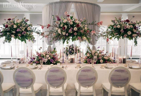 wedding head table with white chairs and large bouquets sitting on the table and propped above with white linen