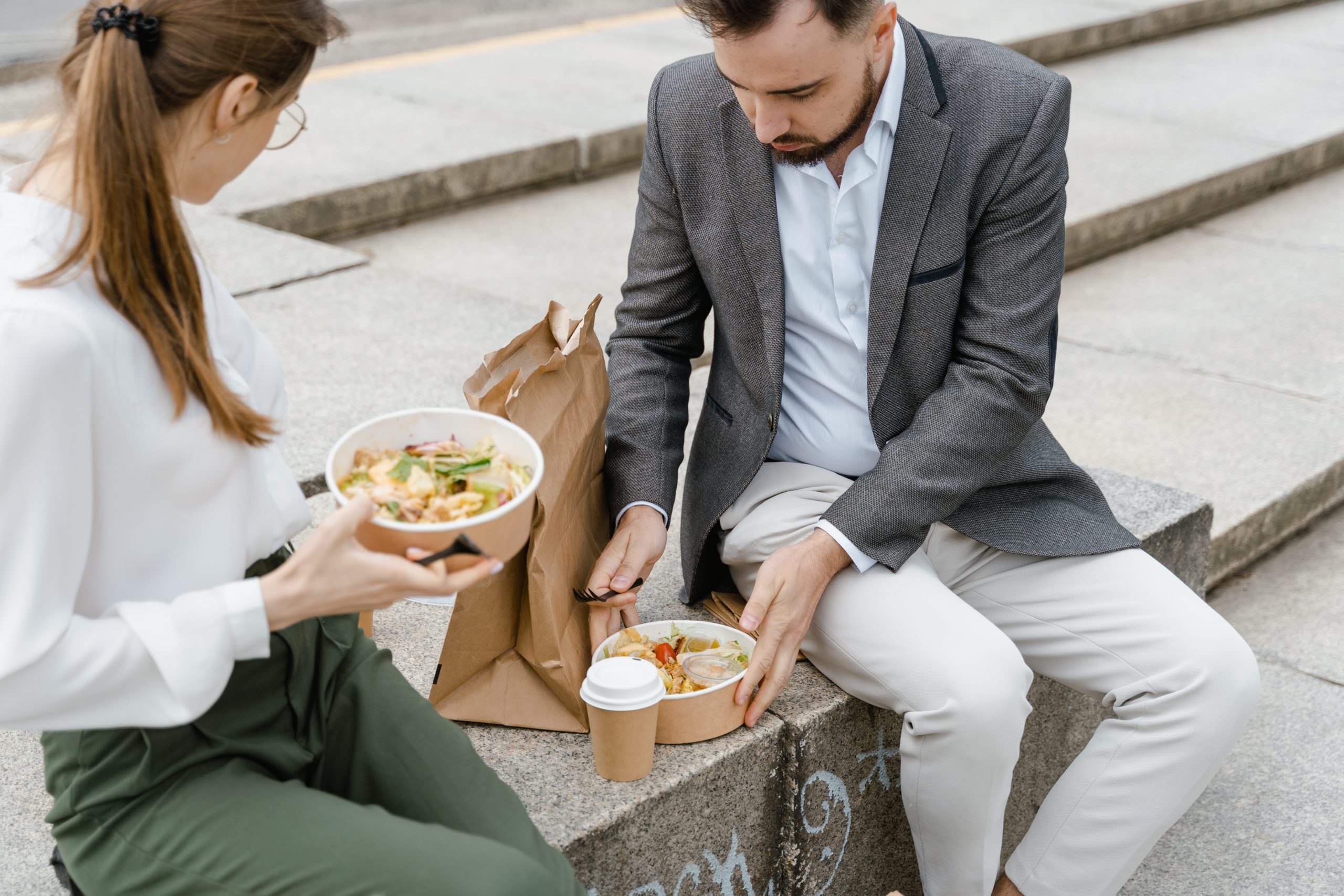 two people eating lunch from takeout containers on a set of steps in dress clothes