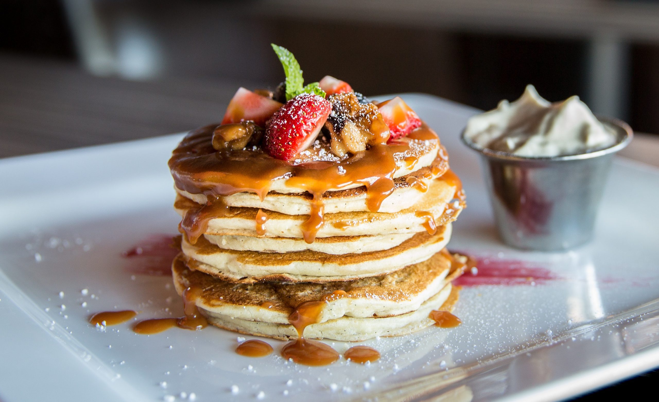 Stack of pancakes with sauce, fruit, and sugar on top with a side of whipped cream