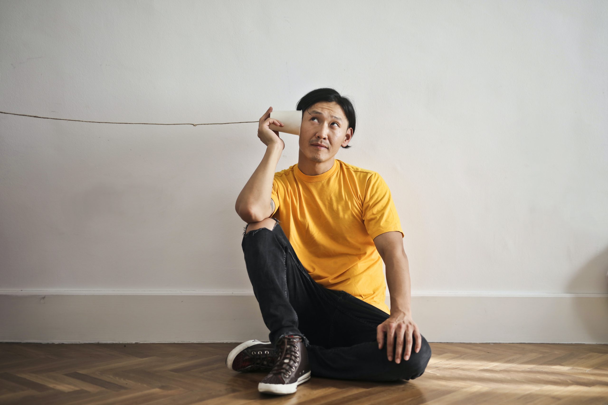 Man with cup to ear with string attached as if listening sitting on floor against the wall