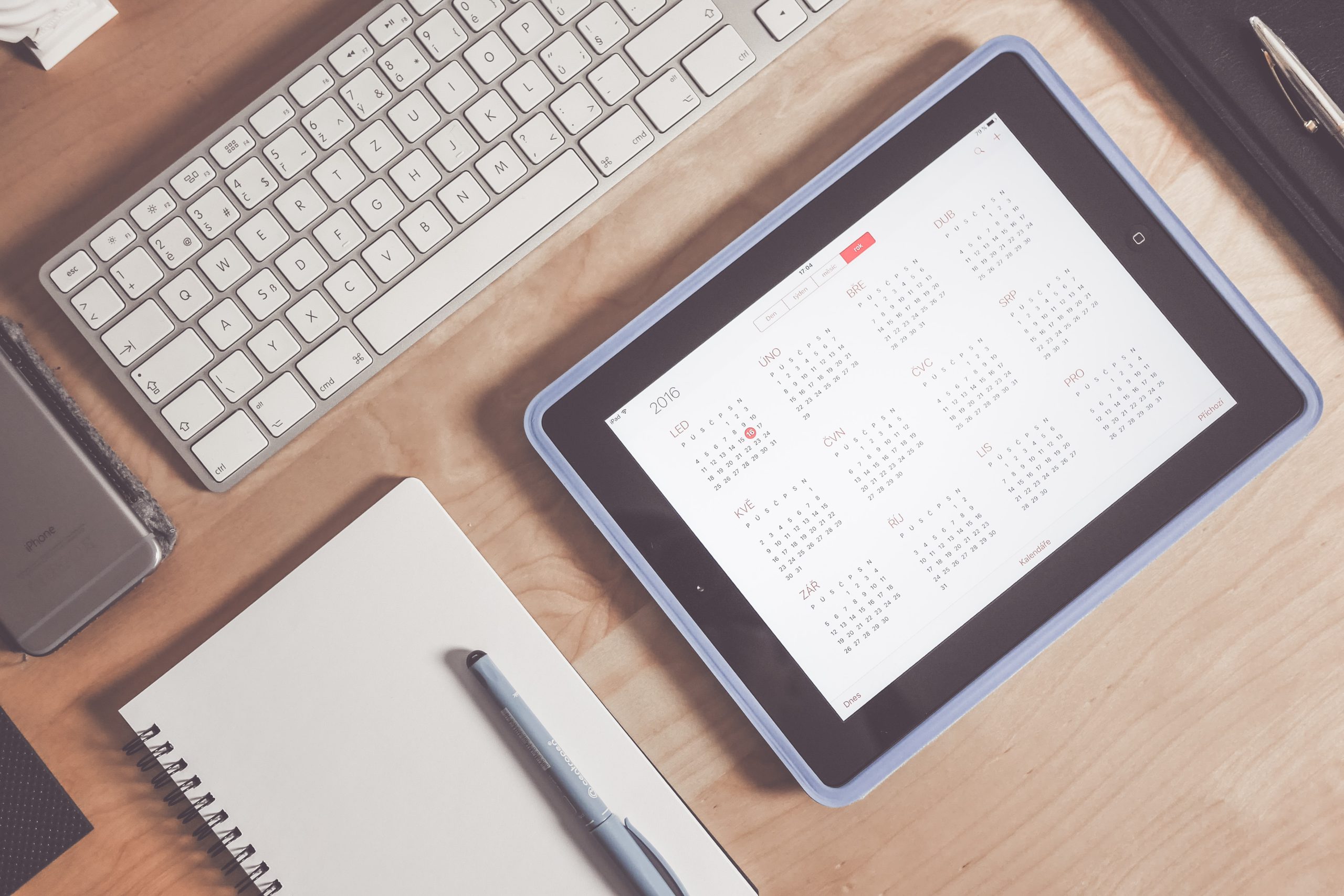 Calendar open on an iPad amongst a notepad, keyboard, and pen on a desk