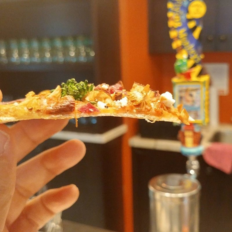 hand holding up thin crust pizza with broccoli, tomatoes, and cheese