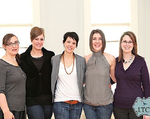 5 woman smiling at the camera linking arms in front of window