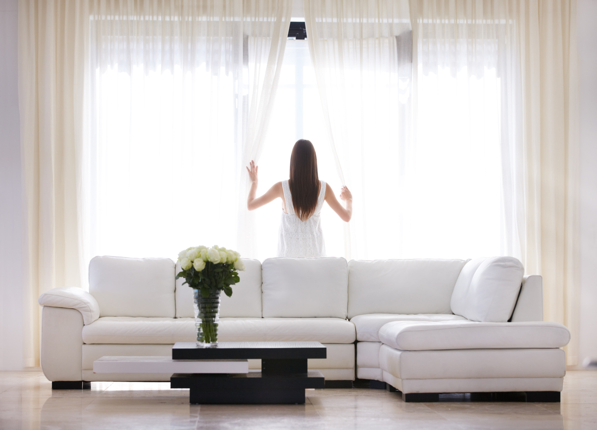 woman in living room behind white couch opening white sheer drapes for window