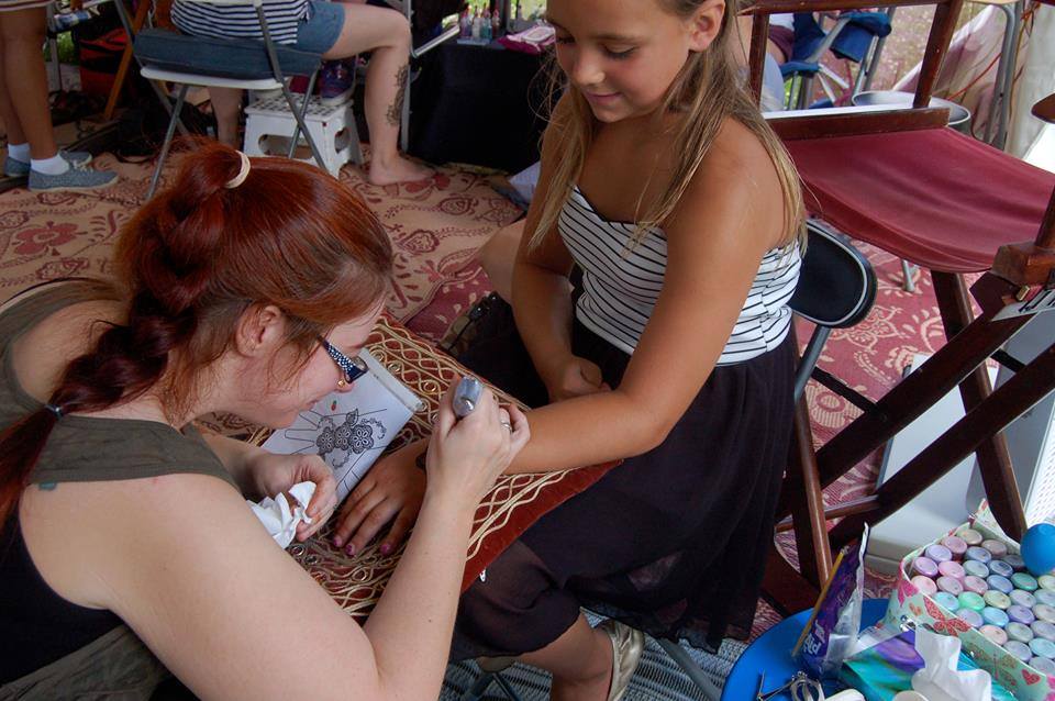red haired woman sitting putting henna tattoo on younger girl sitting down looking happy 