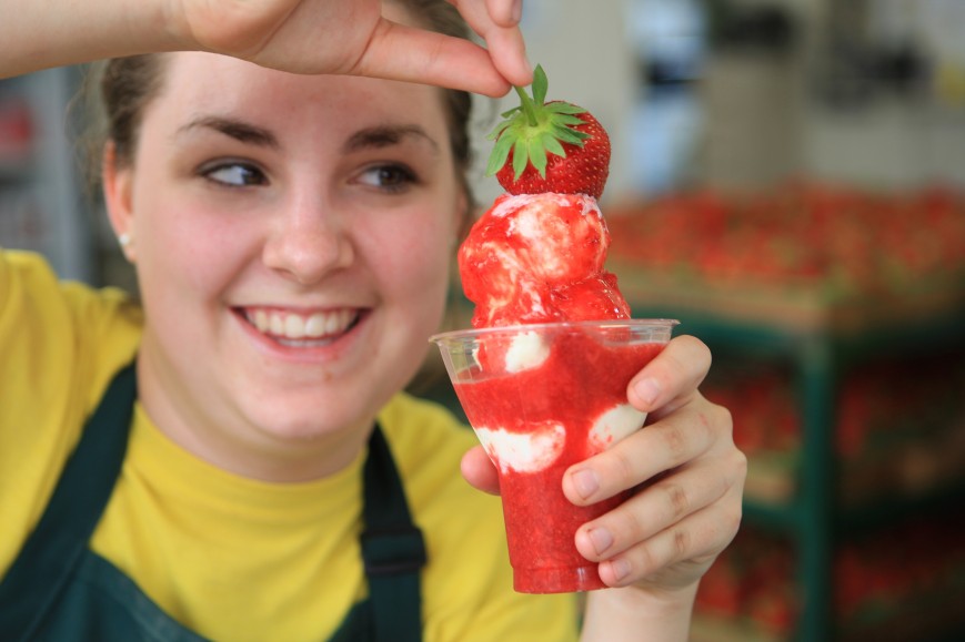 closeup of woman dipping strawberries into a sundae wearing an apron smiling