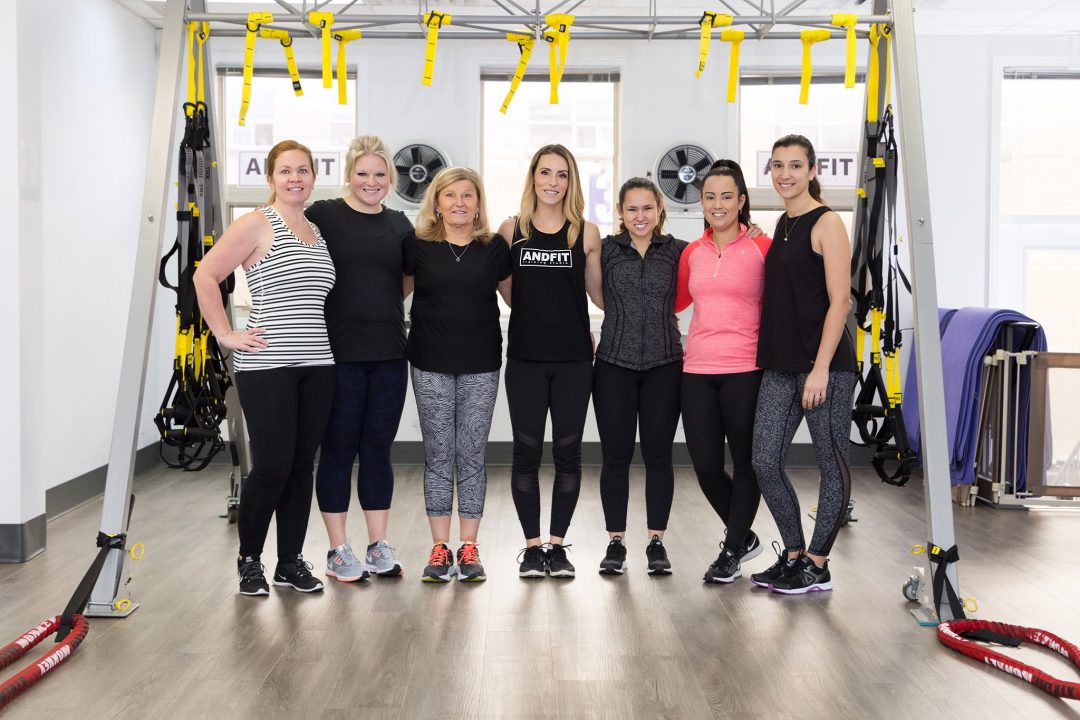 group of women inside fitness centre surrounded by workout equipment linking arms and smiling