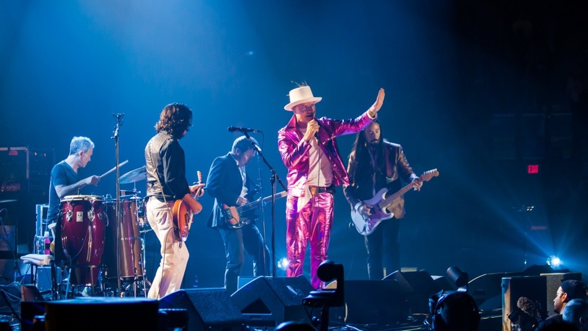 crowd view of band on stage with brightly dressed front man wearing hat waving at audience