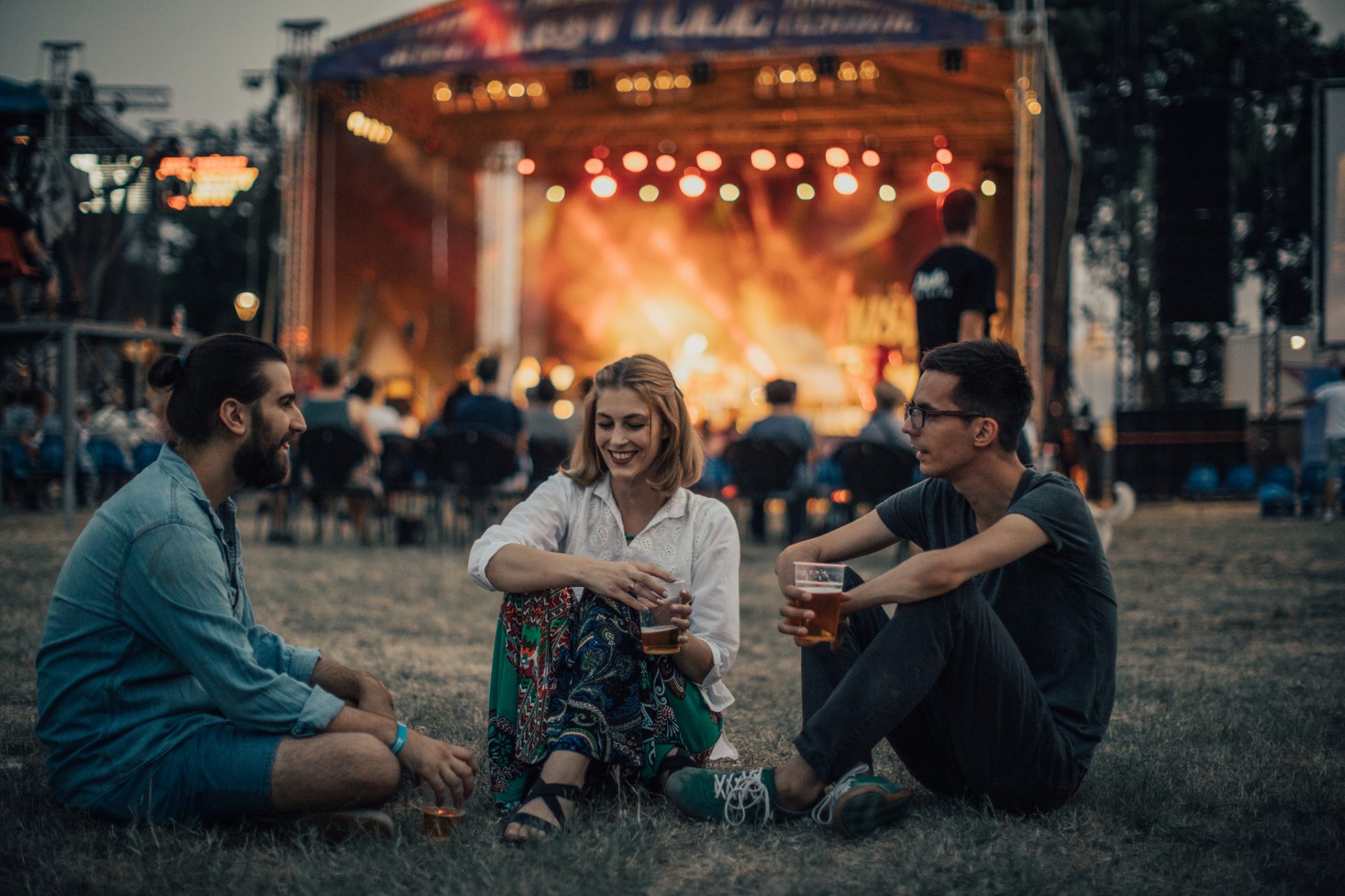 three young adults sitting in front of stage laughing and holding beers with lights in background
