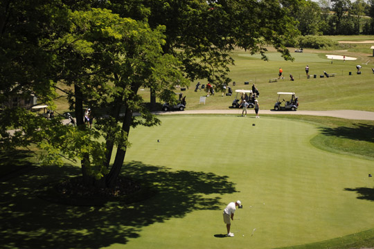 Men on putting green at golf course with carts and other people behind