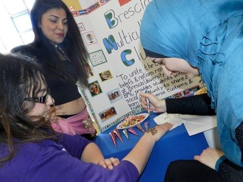 woman with hijab performing henna on younger girl in front of a poster board with another woman looking over