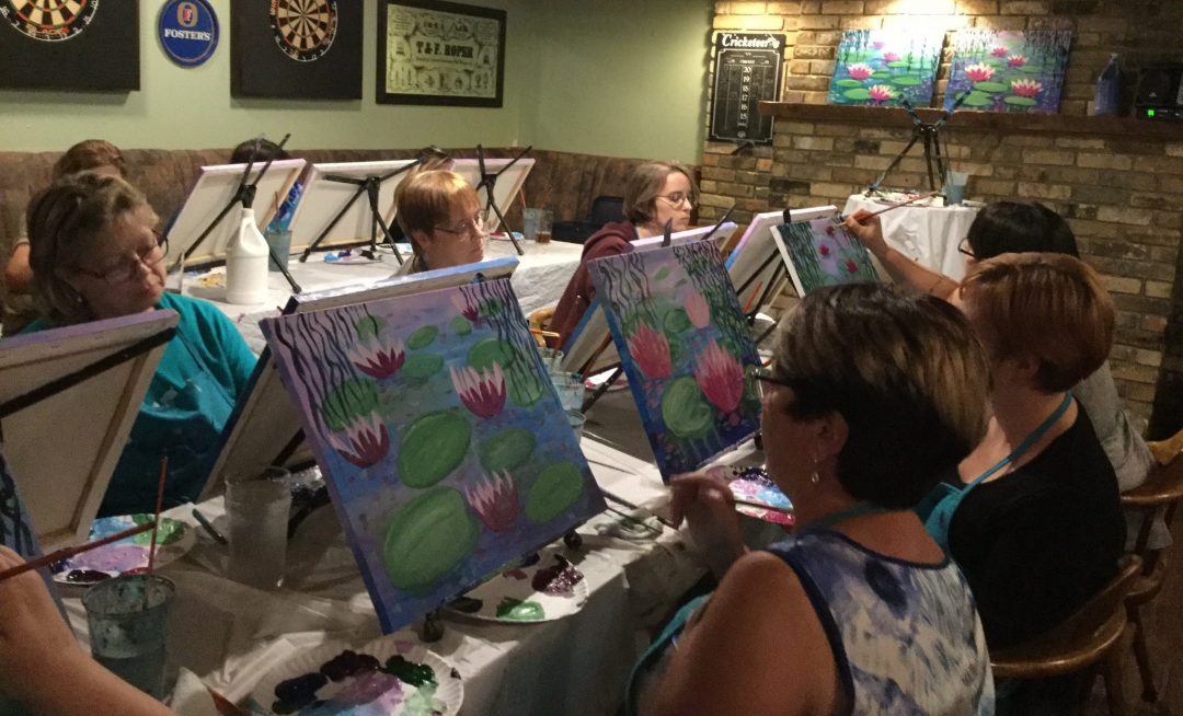 group of women sitting at table each with a small canvas in front with lily pad paintings almost complete
