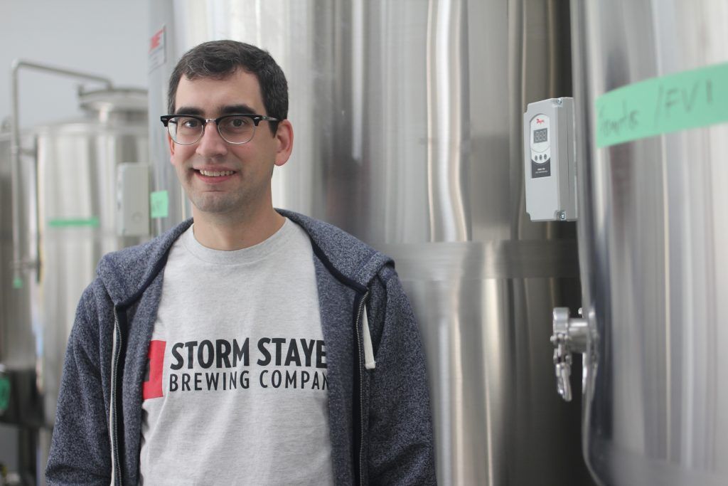close of up brewery employee wearing branded shirt in front of three vats of beer