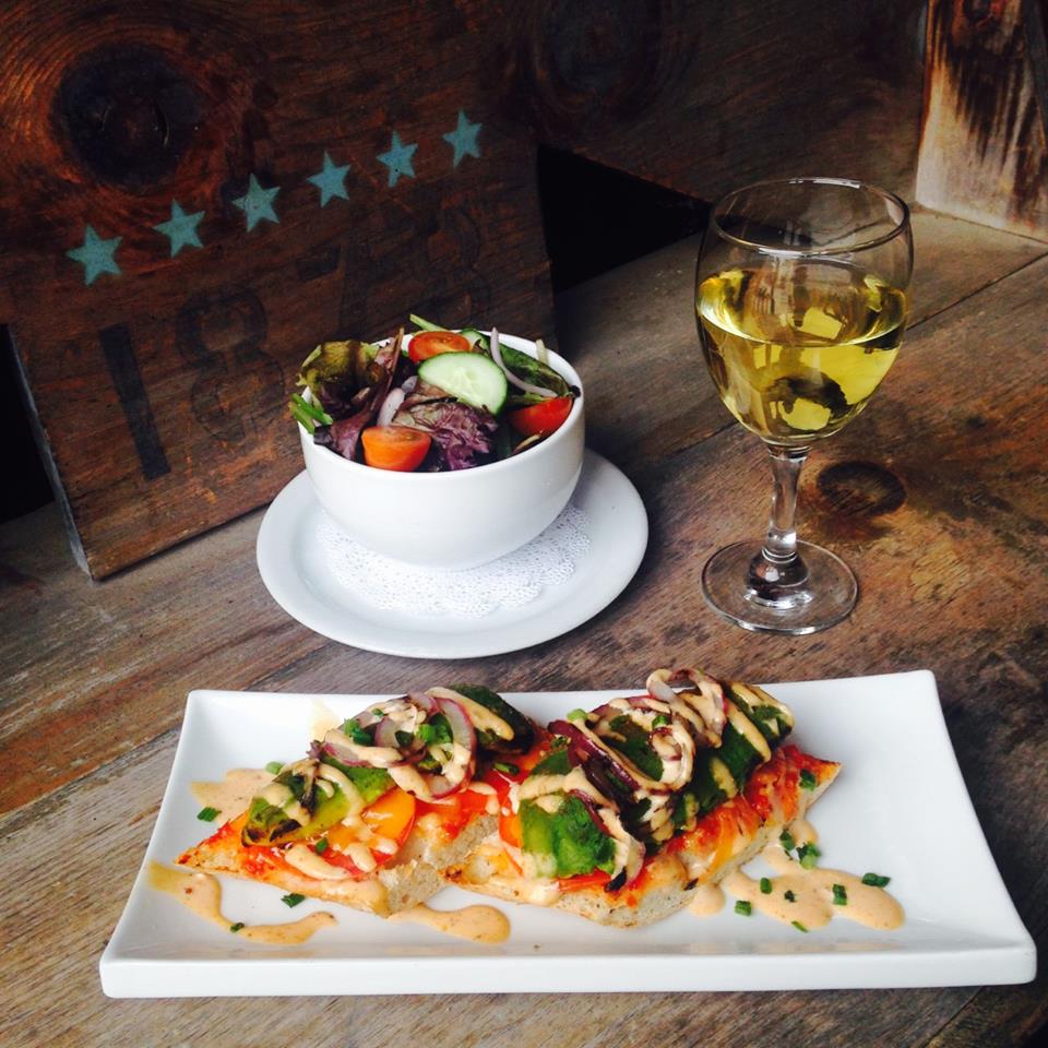 open face sandwich with multi coloured vegetables and an orange drizzle with a bowl of salad on the side and glass of white wine on a wooden rustic table with menu in background