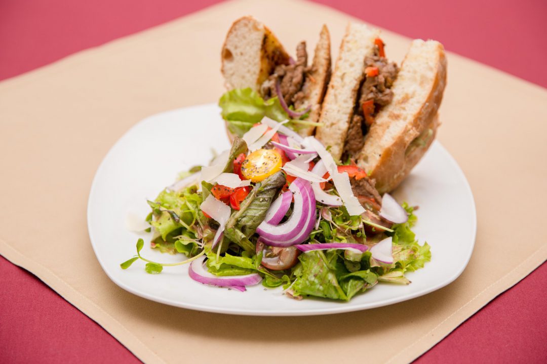 Steak Sandwich on a Baguette with Peppers and onions with a side garden salad on a red table with brown place setting