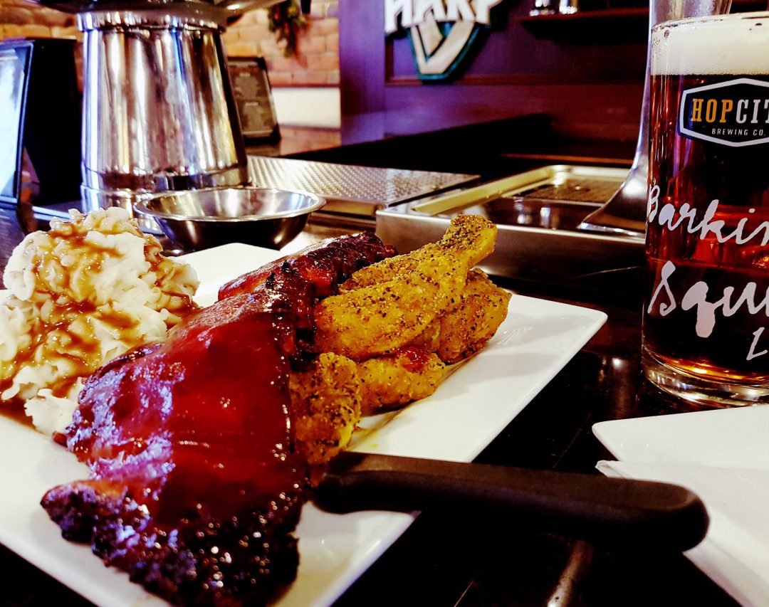 square plate with ribs, mashed potatoes and a red coloured beer in the background in front of the bar