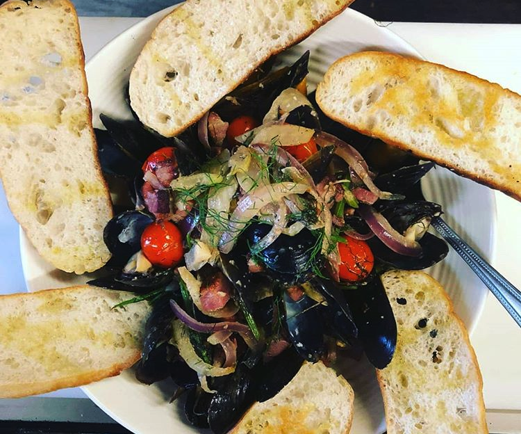 bowl of mussels in shells under multi coloured vegetables with bread surrounding bowl